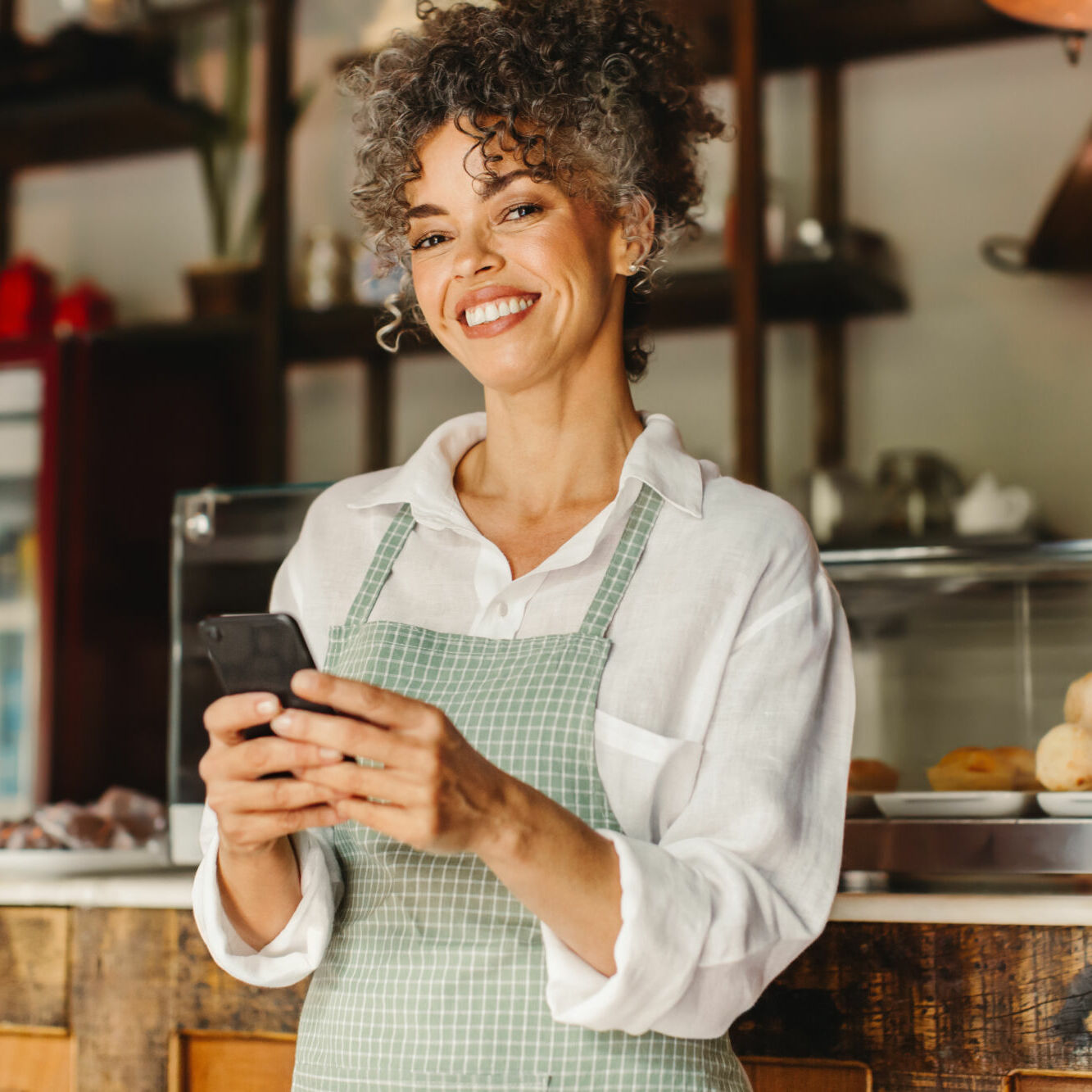 Small business owner using a smartphone in her cafe. Mature businesswoman smiling at the camera while standing in front of the counter. Successful entrepreneur communicating with her customers online.