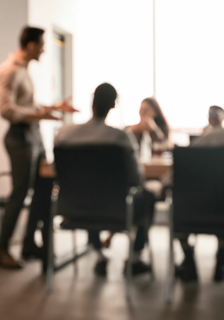 Business Presentation, Blurred Background. Businessman Giving Speech During Seminar With Coworkers In Office, Standing At Desk In Boardroom, Diverse People Sitting At Table And Listening To Speaker