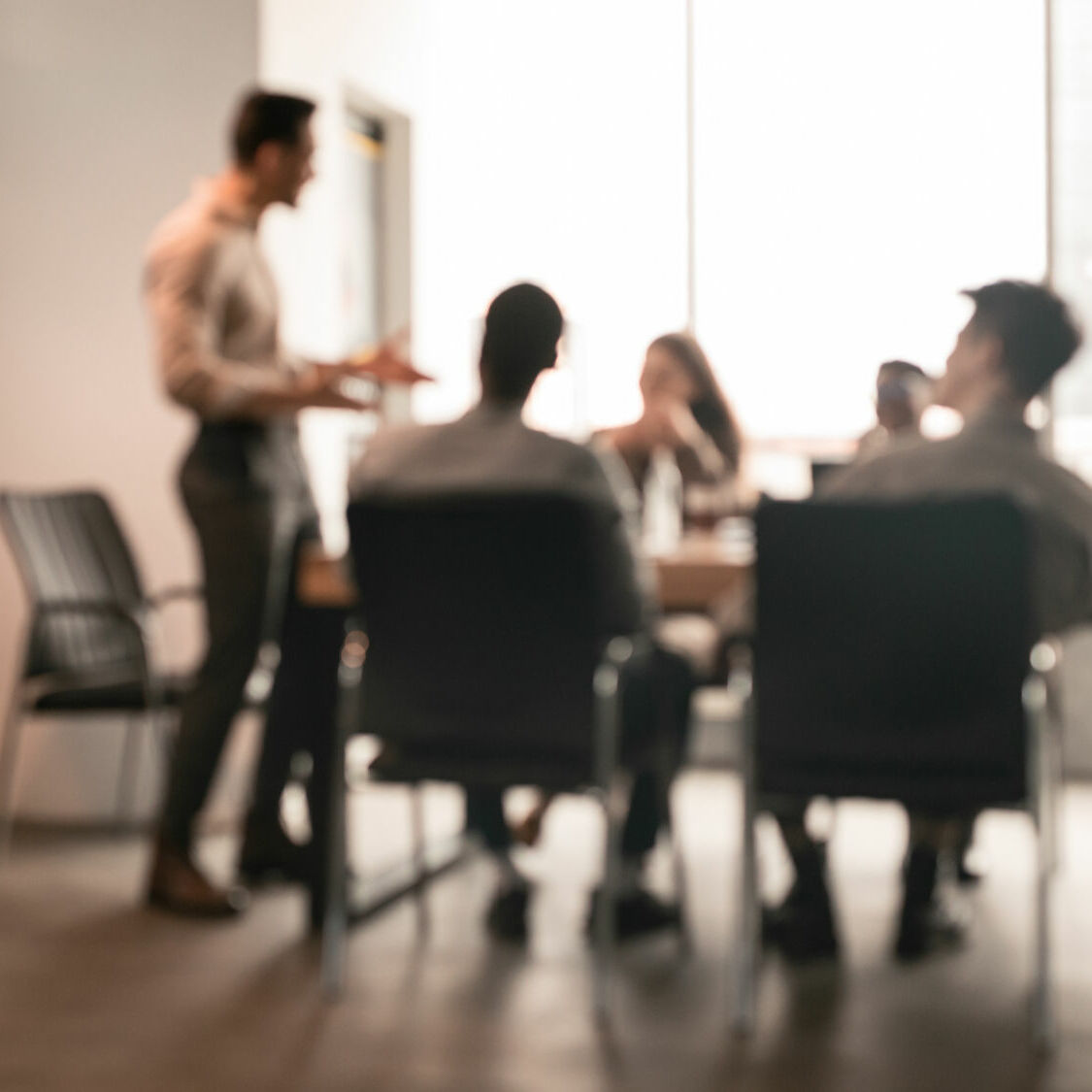 Business Presentation, Blurred Background. Businessman Giving Speech During Seminar With Coworkers In Office, Standing At Desk In Boardroom, Diverse People Sitting At Table And Listening To Speaker